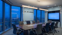 Modern corporate boardroom with panoramic city views through floor-to-ceiling windows at dusk. Executive conference room features sleek table, ergonomic chairs, wall-mounted display.