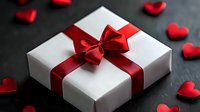 White gift box with red satin ribbon and bow surrounded by small red hearts on dark surface for Valentine's Day, anniversary, romance celebration.