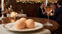 Elegant dinner setting with gourmet dumplings on white plate and rose wine glass in upscale restaurant with warm bokeh lighting and diners in background.