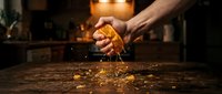 Hand squeezing fresh orange over wooden surface with juice dripping down, warm kitchen lighting in background, citrus preparation for cooking or beverage making.