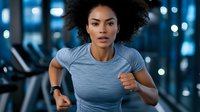 Young African American woman with natural curly hair running on treadmill in modern gym. Athletic female in grey t-shirt during cardio workout with determined expression.