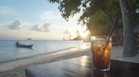Refreshing cola drink with ice on wooden table at sunset beach, fishing boat and palm trees silhouette in background, tropical vacation atmosphere.