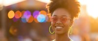 Young African American woman with curly afro hair and glasses smiling joyfully in warm golden hour sunlight with colorful bokeh lights in background.