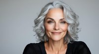 Confident mature woman with natural gray wavy hair wearing black top smiling at camera against neutral background, radiating positive aging and self-acceptance.