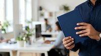 Man in dark navy button-up shirt holding blue tablet device in modern office workspace with plants and natural light for business technology concepts.