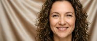 Smiling middle-aged Caucasian woman with curly brown hair against beige fabric background. Professional headshot for business, healthcare, or lifestyle content.