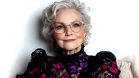 Elegant senior Caucasian woman with silver curly hair wearing stylish glasses and floral black dress, smiling confidently in professional portrait.