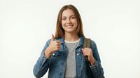 Young woman with long brown hair and fair complexion wearing denim jacket over striped shirt, carrying backpack, smiling at camera with thumbs up gesture for education, travel, or lifestyle content.