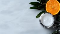 Fresh orange slice and natural cream moisturizer on gray background with green leaves and copy space for skincare and cosmetic product presentation.