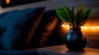 Moody evening interior with black textured vase holding green leaves against warm fairy lights and dark blue pillows, creating cozy atmospheric home decor setting.