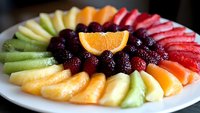 Colorful fresh fruit platter with sliced kiwi, pineapple, orange, strawberries and mixed berries arranged in rainbow pattern on white plate, food photography.