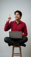 Young Indian man in red shirt pointing up with eureka moment while using laptop on wooden stool against gray background for business concepts.
