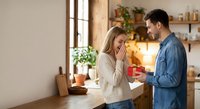 Young Caucasian man surprising blonde woman with red gift box in modern kitchen during romantic moment together at home.