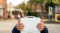 Young child with medium skin tone holding white lunch box or storage case in front of face at outdoor playground, suitable for education, childhood, and product mockup concepts.