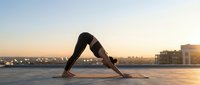 Woman practicing downward facing dog yoga pose on rooftop at sunset with city skyline background for wellness and fitness lifestyle content.