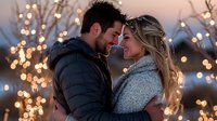 Romantic couple embracing outdoors on winter evening with glowing fairy lights creating magical bokeh atmosphere in background, showing intimate moment.