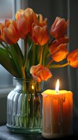 Vibrant orange tulips arranged in clear glass vase with lit candle, creating warm spring ambiance for home decor and seasonal celebrations.