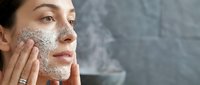Young woman applying exfoliating scrub mask to face during skincare routine in modern bathroom for healthy glowing skin care treatment.