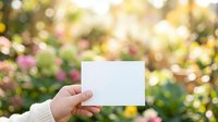 Hand holding blank white card against blooming garden with pink and yellow flowers, ideal for spring promotions, greeting cards, and seasonal marketing campaigns.