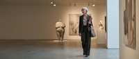 Senior woman with gray hair wearing black formal suit walking through contemporary art gallery with sculptures and paintings displayed on white walls.