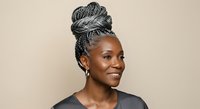 Smiling African American woman with gray braided updo hairstyle wearing black shirt against neutral beige background, representing mature beauty and natural hair styling.