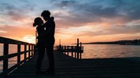 Silhouette of man holding woman on wooden pier at sunset over calm water, romantic couple embracing during golden hour by lake or sea.