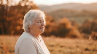 Senior Caucasian woman with gray hair smiling peacefully outdoors during golden hour sunset with warm autumn landscape and mountains in background.