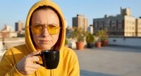Young Caucasian woman in yellow hoodie and sunglasses drinking coffee on urban rooftop terrace with city skyline background during golden hour morning routine.
