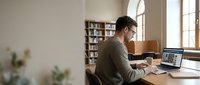 Man in olive sweater working at desk with laptop in bright library study room with bookshelves and natural window light, suitable for education, remote work, and productivity concepts.