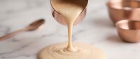 Creamy caramel sauce pouring from copper ladle creating perfect spiral on marble surface with copper cookware background for culinary and dessert preparation.