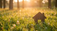 Small wooden house model sitting in green grass field with warm golden sunlight creating bokeh effect for real estate and home ownership concepts.