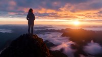 Young woman silhouetted on mountain peak at sunrise, overlooking dramatic cloudscape with golden sun breaking through clouds and misty valleys below.