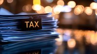 Stack of paper documents labeled tax with blurred bokeh lights in background, dramatic lighting creates moody atmosphere for financial concept.