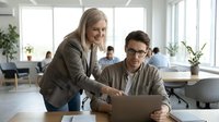 Professional businesswoman mentor coaching young male colleague at laptop in modern bright office workspace for corporate training and development.