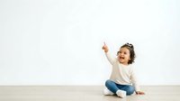 Young child with curly dark hair, sitting on wooden floor against white wall, pointing upward with expression of wonder and discovery, wearing casual cream and denim clothing for educational and parenting concepts.