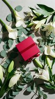 Red gift box surrounded by white lilies and eucalyptus leaves on neutral background for holiday celebrations and special occasions.
