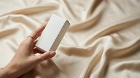 White rectangular box mockup held in hand against beige fabric background for product packaging design and branding presentations.