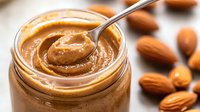 Creamy almond butter in glass jar with metal spoon, surrounded by fresh whole almonds on wooden surface. Natural organic spread with smooth texture.