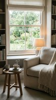 Cozy reading nook with comfortable armchair, wooden side table, books and coffee cup by sunny window overlooking lush garden for relaxation and leisure.