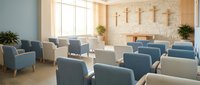 Modern church interior with blue upholstered chairs arranged in rows facing wooden cross on brick wall. Natural light streams through large windows.