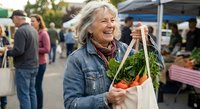Happy senior Caucasian woman with gray hair holding canvas bag with fresh vegetables at outdoor farmers market, smiling joyfully while shopping for organic produce.