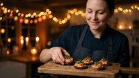 Female chef in black uniform garnishing gourmet appetizers on wooden board in restaurant kitchen with warm bokeh lighting for culinary presentation.