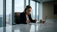 Young professional woman with long blonde hair and glasses, wearing dark business attire, working at marble desk in modern high-rise office with city skyline view, using laptop for corporate work.