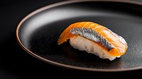 Fresh salmon nigiri sushi with thin slice of raw fish on pressed rice, served on dark ceramic plate against black background, dramatic lighting.