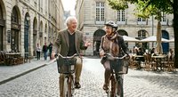 Senior couple riding vintage bicycles through historic cobblestone street with classic architecture, enjoying active retirement lifestyle together.