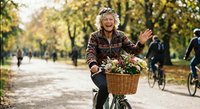 Cheerful mature Caucasian woman cycling through autumn park with flower basket, waving hello while enjoying outdoor recreation and healthy lifestyle.