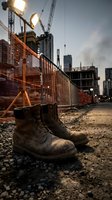 Worn work boots on dusty ground at active construction site with cranes, metal fencing, and city skyline at dusk, representing labor, industrial development, and urban building projects.