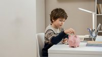 Young boy with light brown hair, Caucasian ethnicity, sitting at desk putting coin into pink piggy bank. Educational finance learning concept for children's savings and money management.