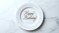 White ceramic plate with Happy Birthday written in chocolate script, decorated with silver sprinkles, on a marble surface. Top view.