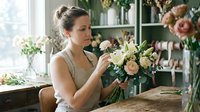 Florist woman arranging fresh flowers in vase at wooden table near window with shelves of blooms and greenery for floral design work.